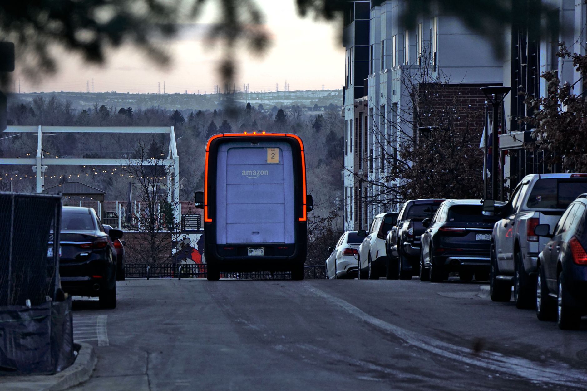 Amazon delivery van driving through an urban street at twilight with cars parked along the side.